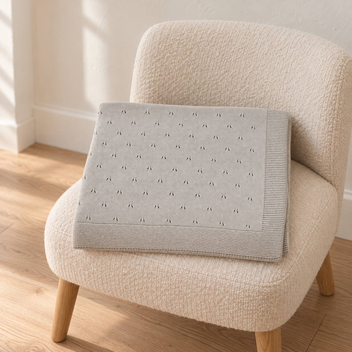 Pislak Zora children’s blanket in grey, neatly folded on a cream upholstered chair seat, showing the tiny repeating eyelet pattern and knitted border.
